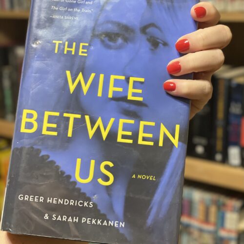 A hand holding the book The Wife Between Us, in front of a bookshelf.