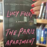 a hand holding the paris apartment book by lucy foley in front of a bookshelf